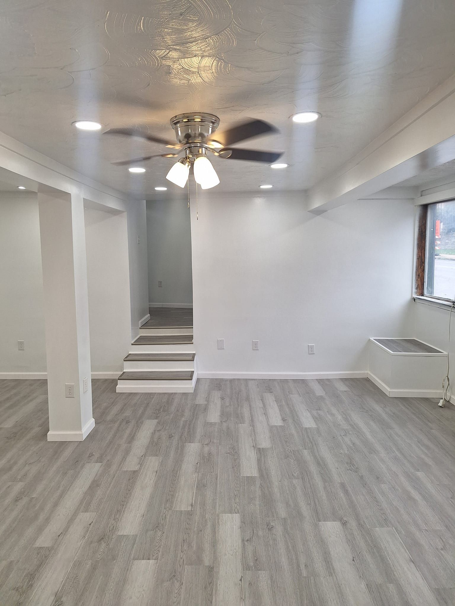 A bright, open-plan room with white walls, recessed lighting, a ceiling fan, gray wood-look flooring, and short stairs.