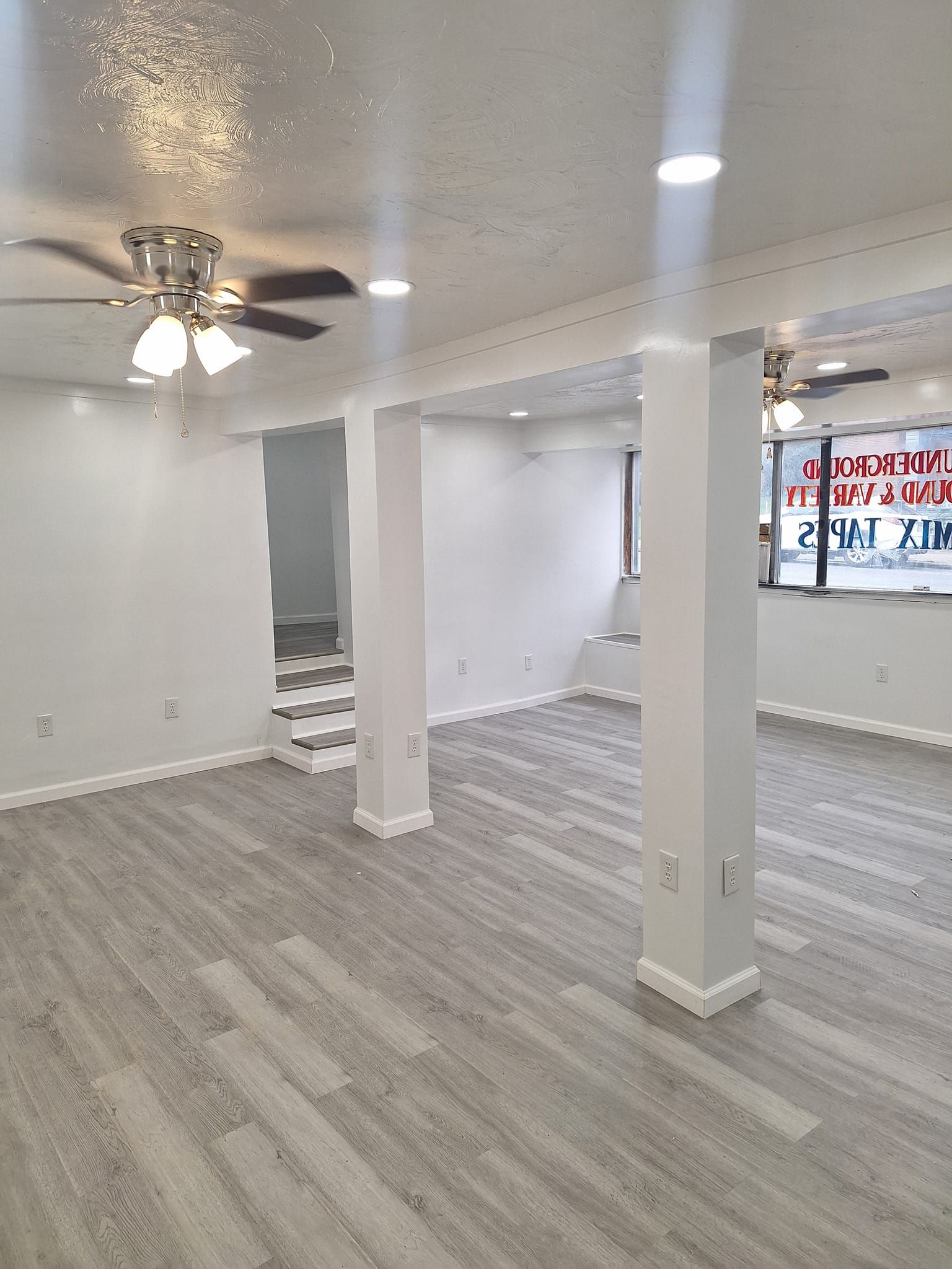 A wide, empty room with light gray wood-look flooring, white walls, support columns, and overhead lighting.