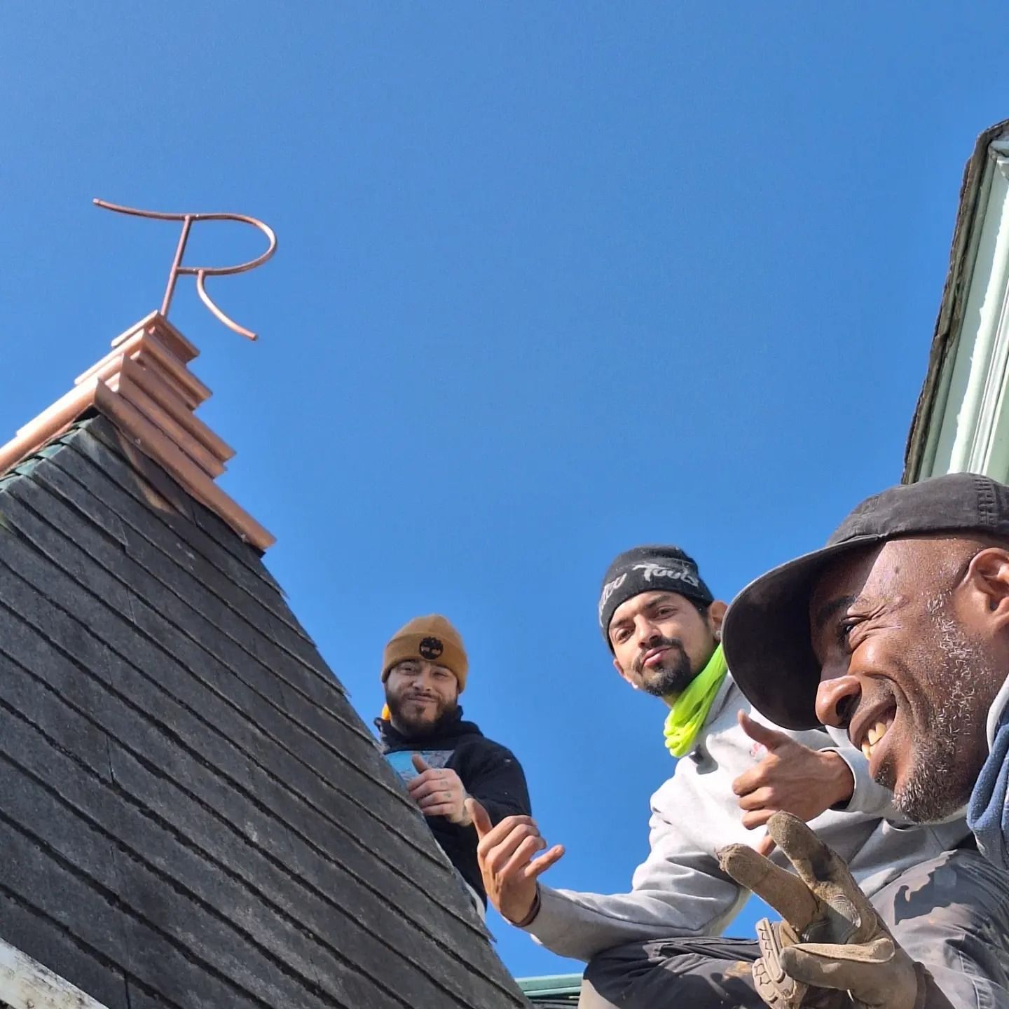 Three people smile and give thumbs-up while standing on a shingled roof next to a copper finial shaped like the letter R.