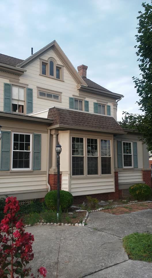 A cream-colored, two-story house with green shutters, a brown roof, and a paved front walkway under a cloudy sky.