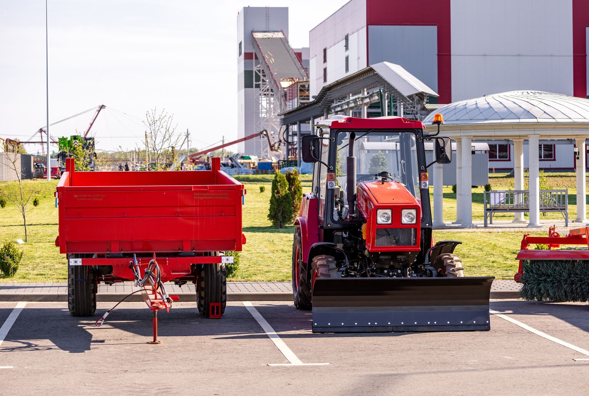 Red tractor with a front blade parked beside a red trailer in an outdoor area near industrial buildings.