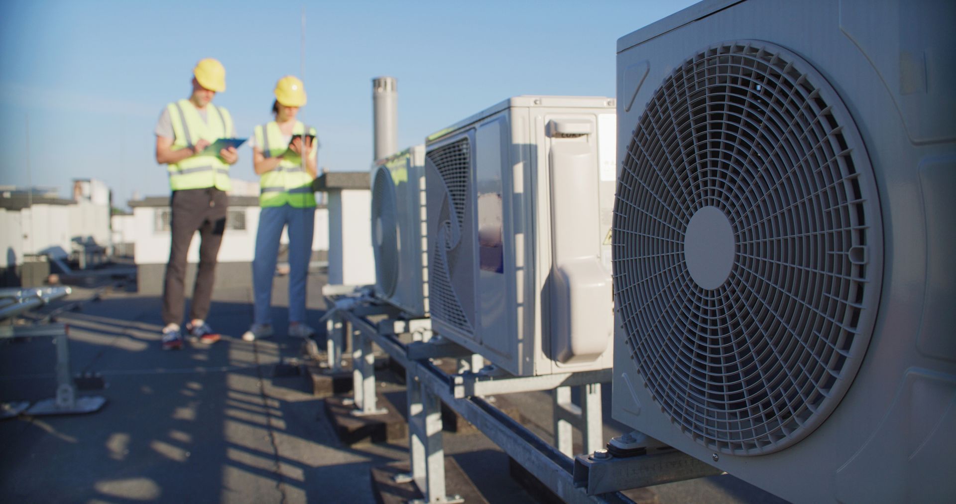 Close-up of a HVAC external unit on top of a building, with blurry engineers on the background.