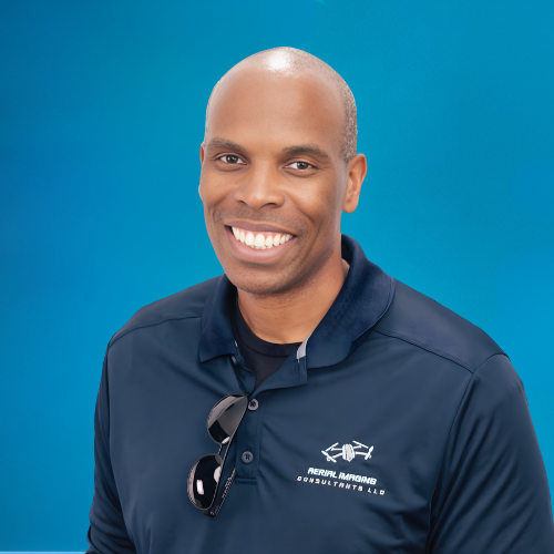 Man with shaved head smiles, wearing a navy polo shirt with Aerial Surveying logo; blue background.