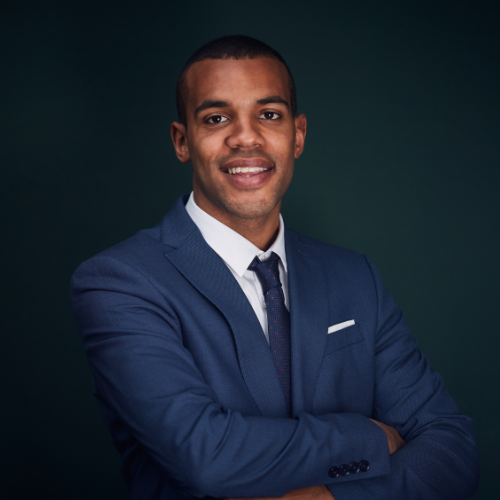 Man in a blue suit, arms crossed, smiling. Dark green background.
