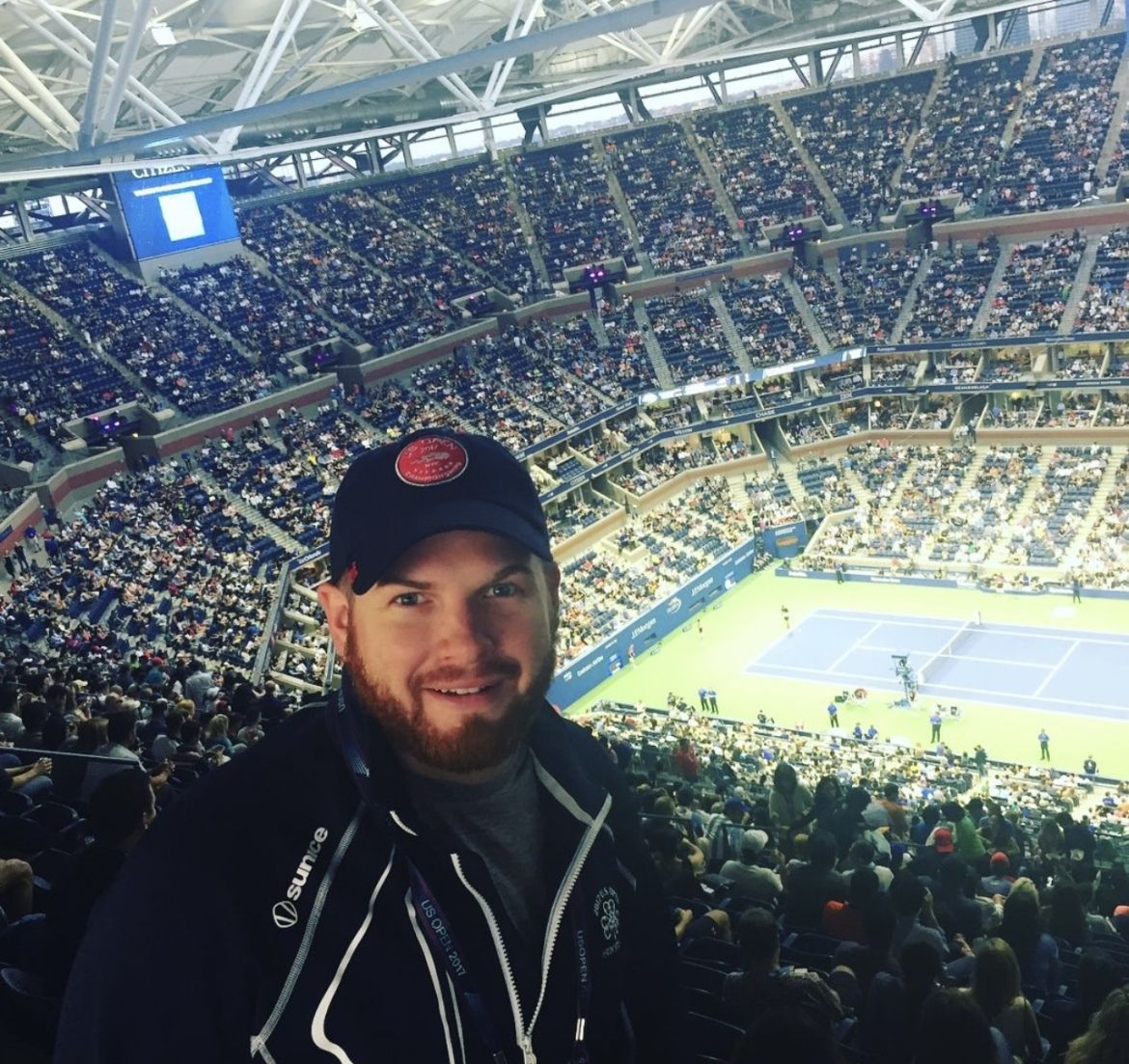 Man in a stadium wearing a baseball cap smiles at camera, tennis court in the background, filled with spectators.