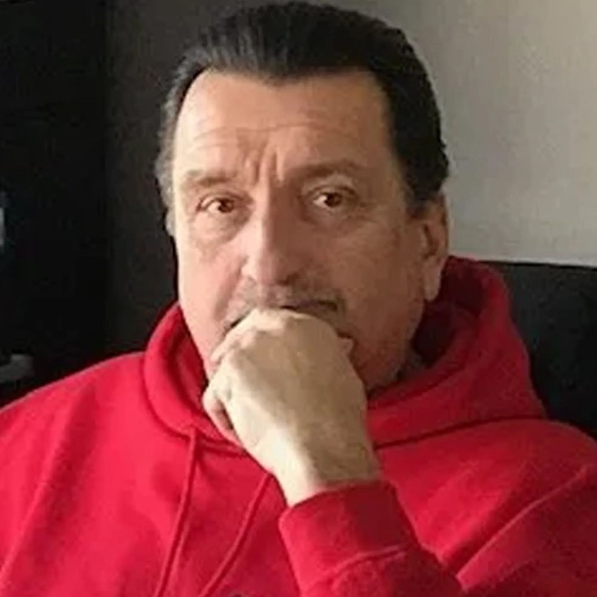 A man in a red hoodie is sitting in a chair with his hand on his chin.