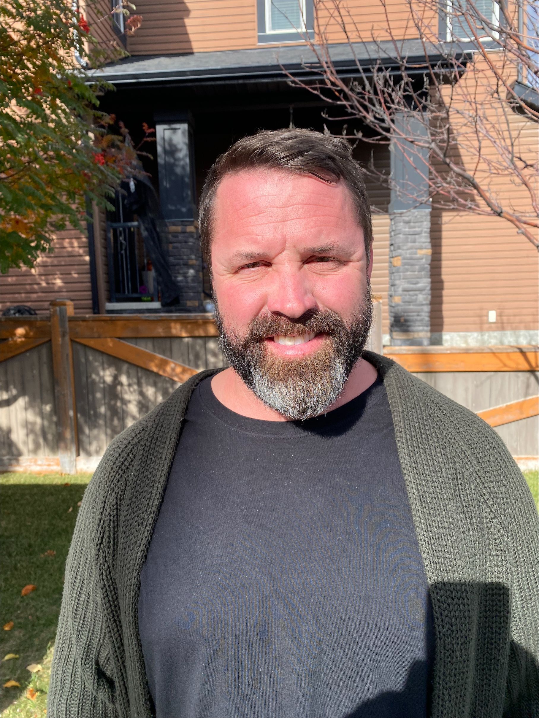 A man with a beard and a green sweater is standing in front of a house.