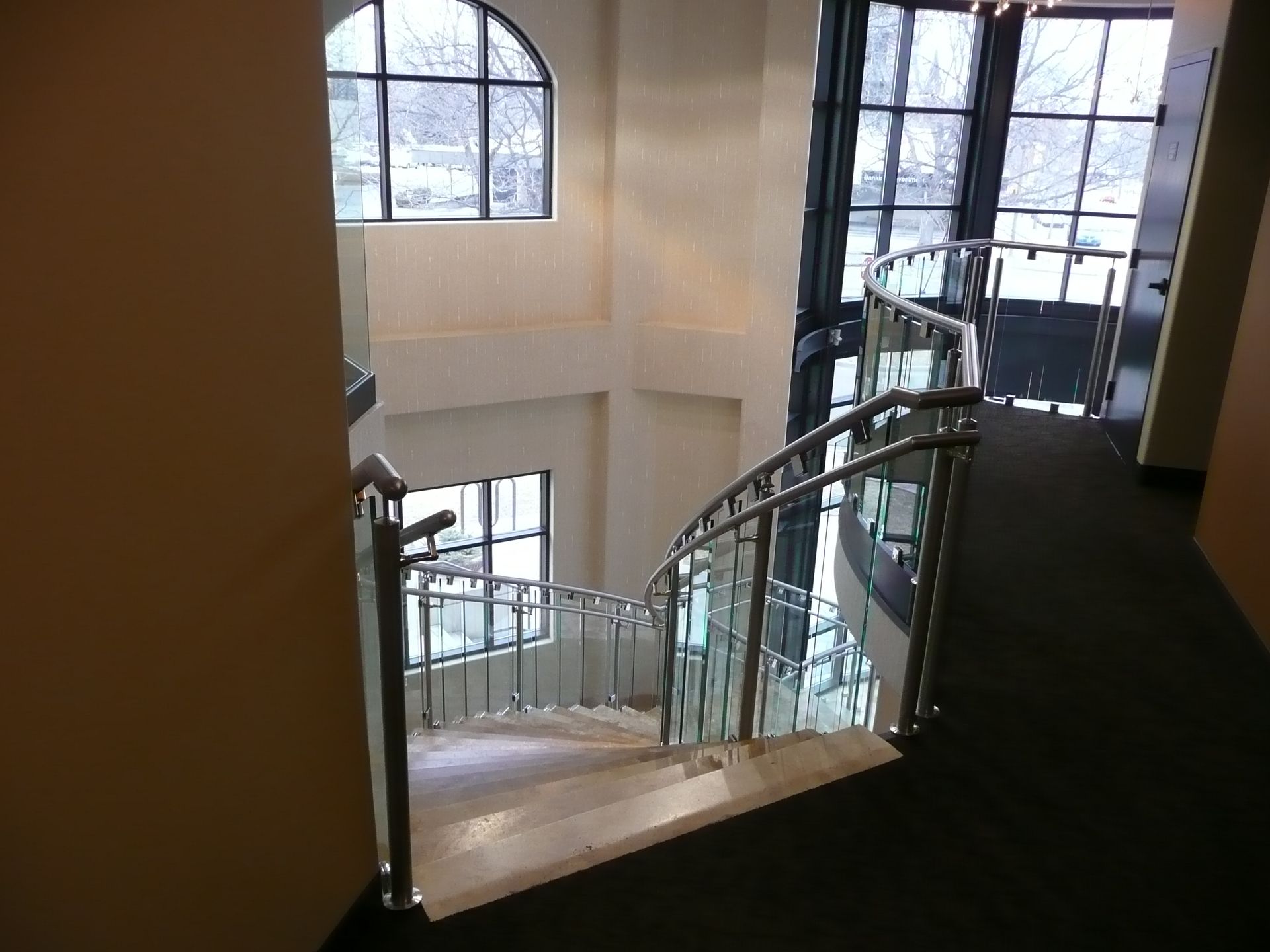 Curved staircase with glass railing in a bright interior lobby beside large arched windows