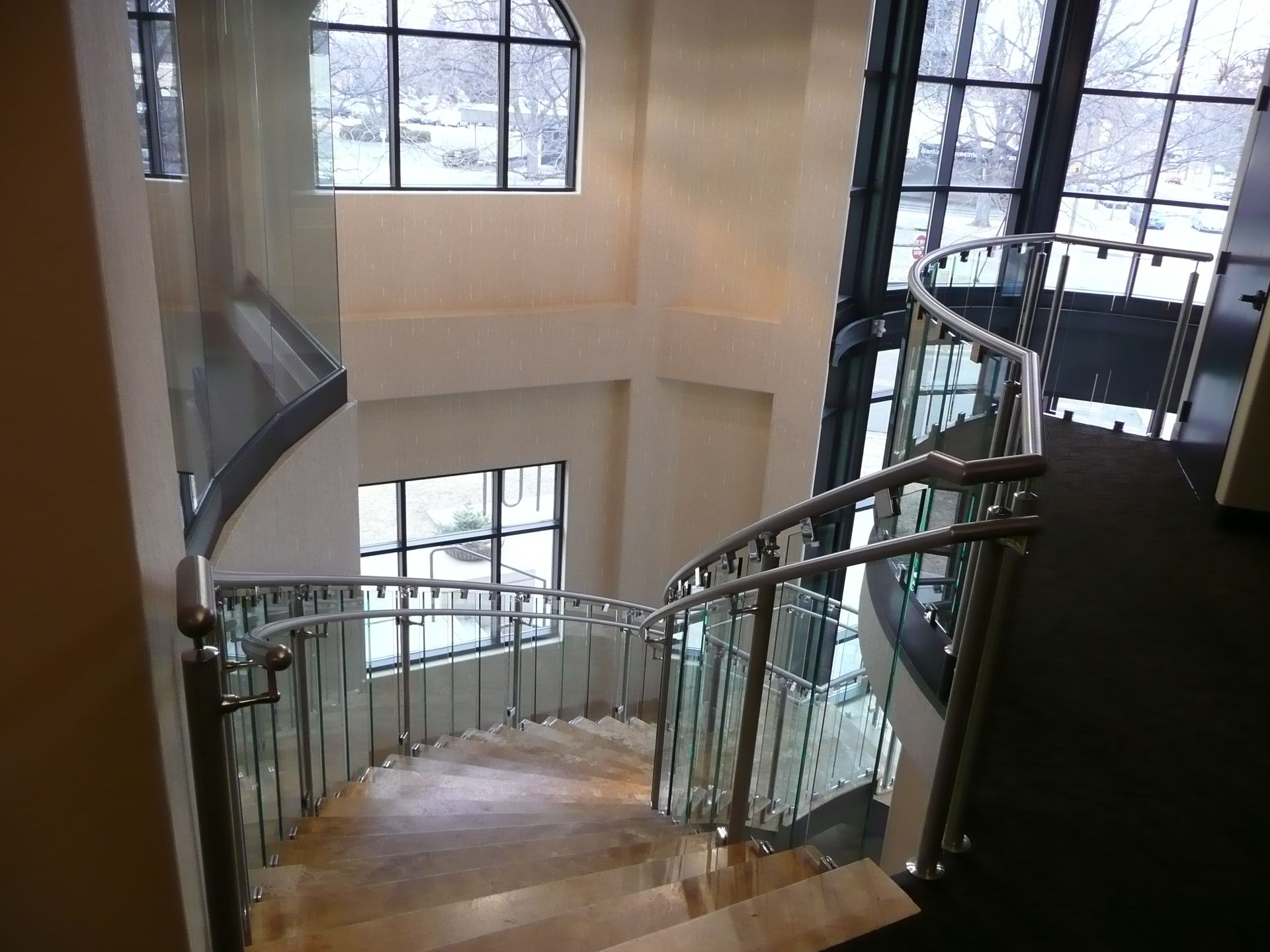 Curved staircase with glass railing in a bright atrium, viewed from the landing above.