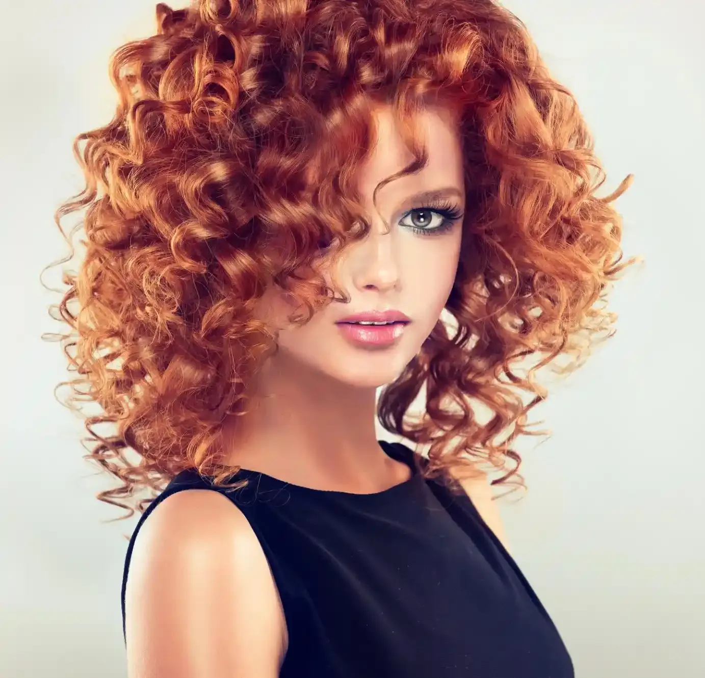 Una donna con i capelli rossi e ricci, che indossa un top nero, posa su uno sfondo chiaro.