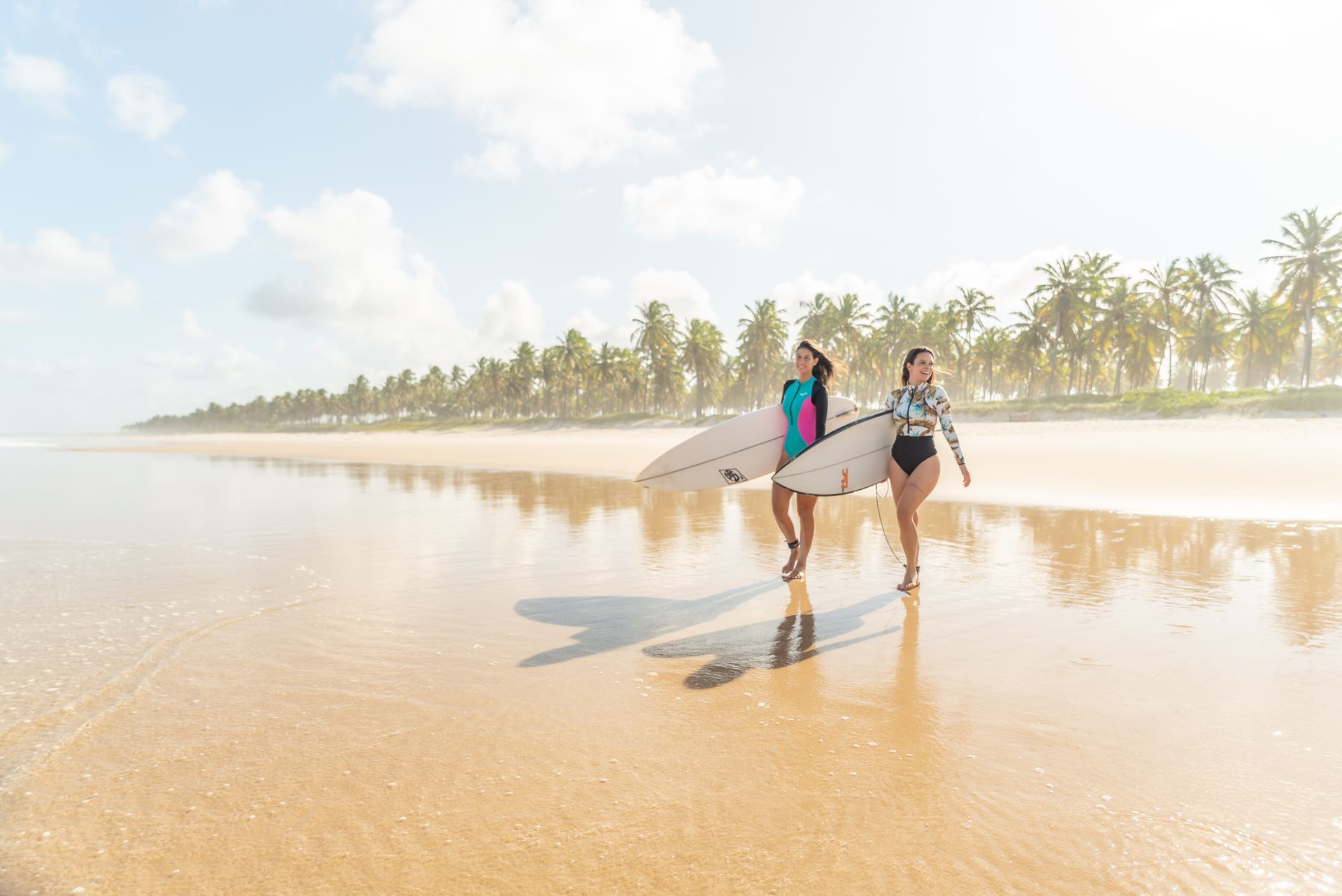 Duas muleres com pranchas de surf na paisagem delumbrante dos coqueirais e mar da Praia do Francês.