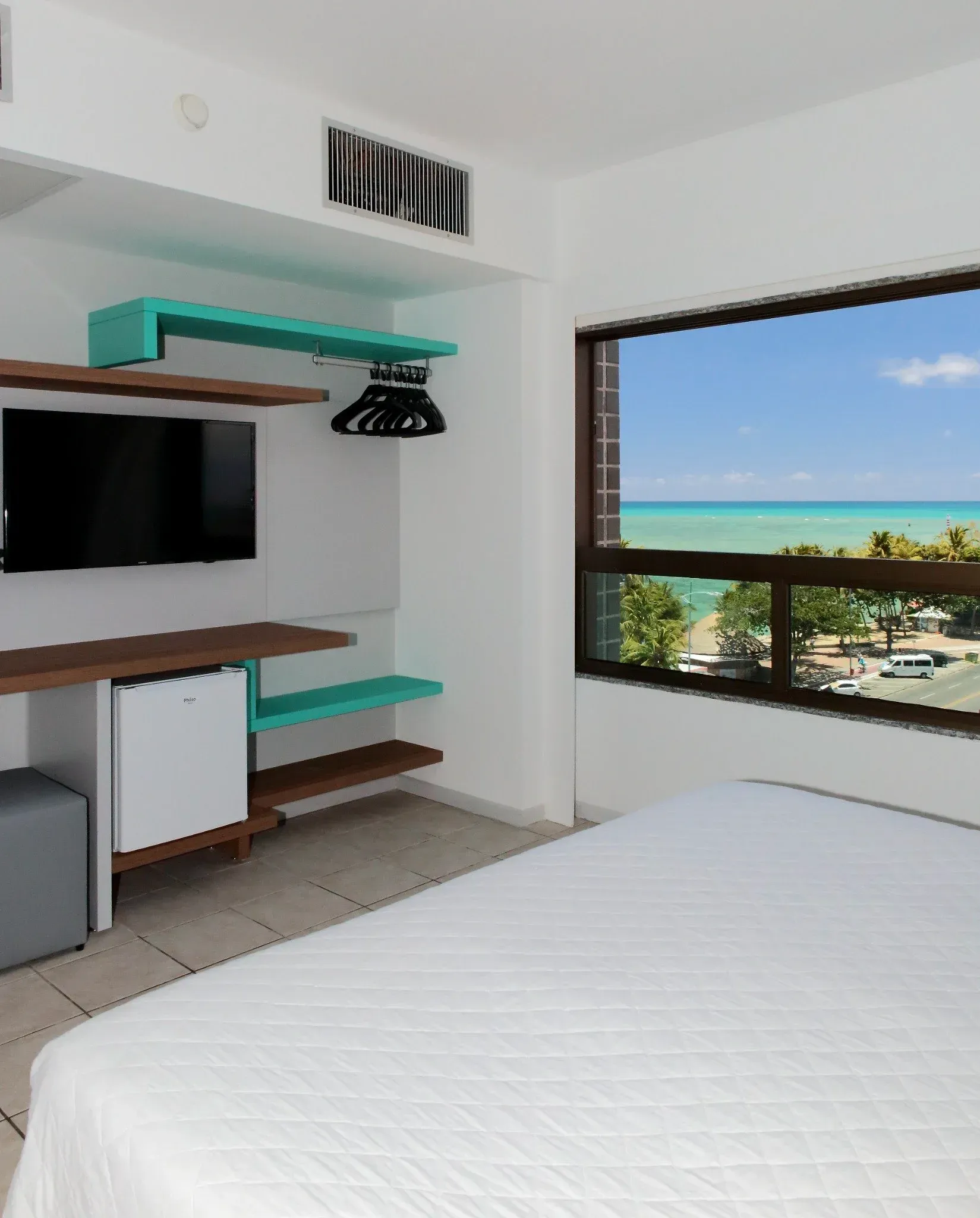 Um quarto de hotel com uma cama, televisão, prateleiras e uma janela com vista para o oceano.