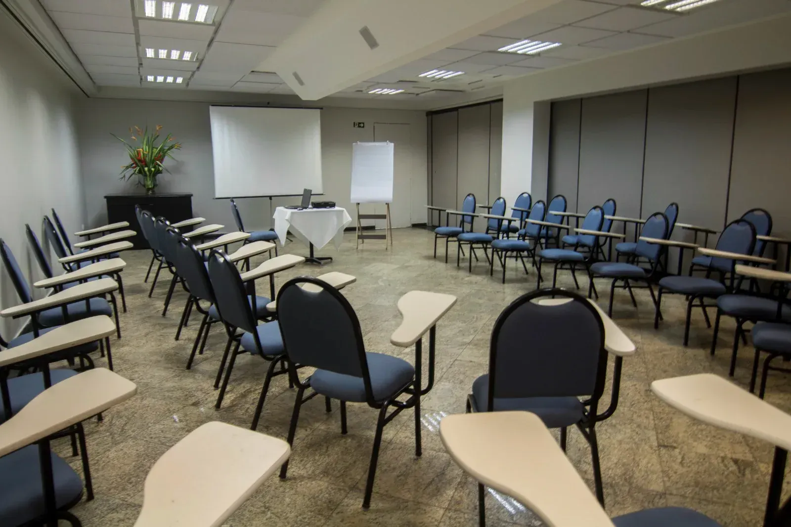 Uma sala de conferências com fileiras de cadeiras e mesas e uma tela de projetor.