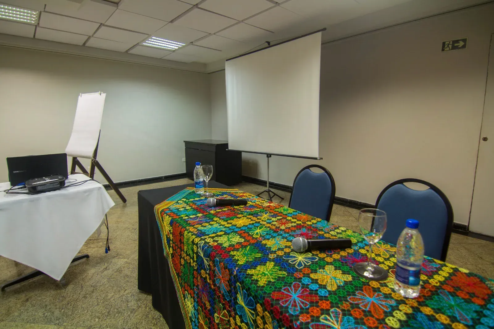 Uma sala de conferências com uma mesa, cadeiras e uma tela de projetor.