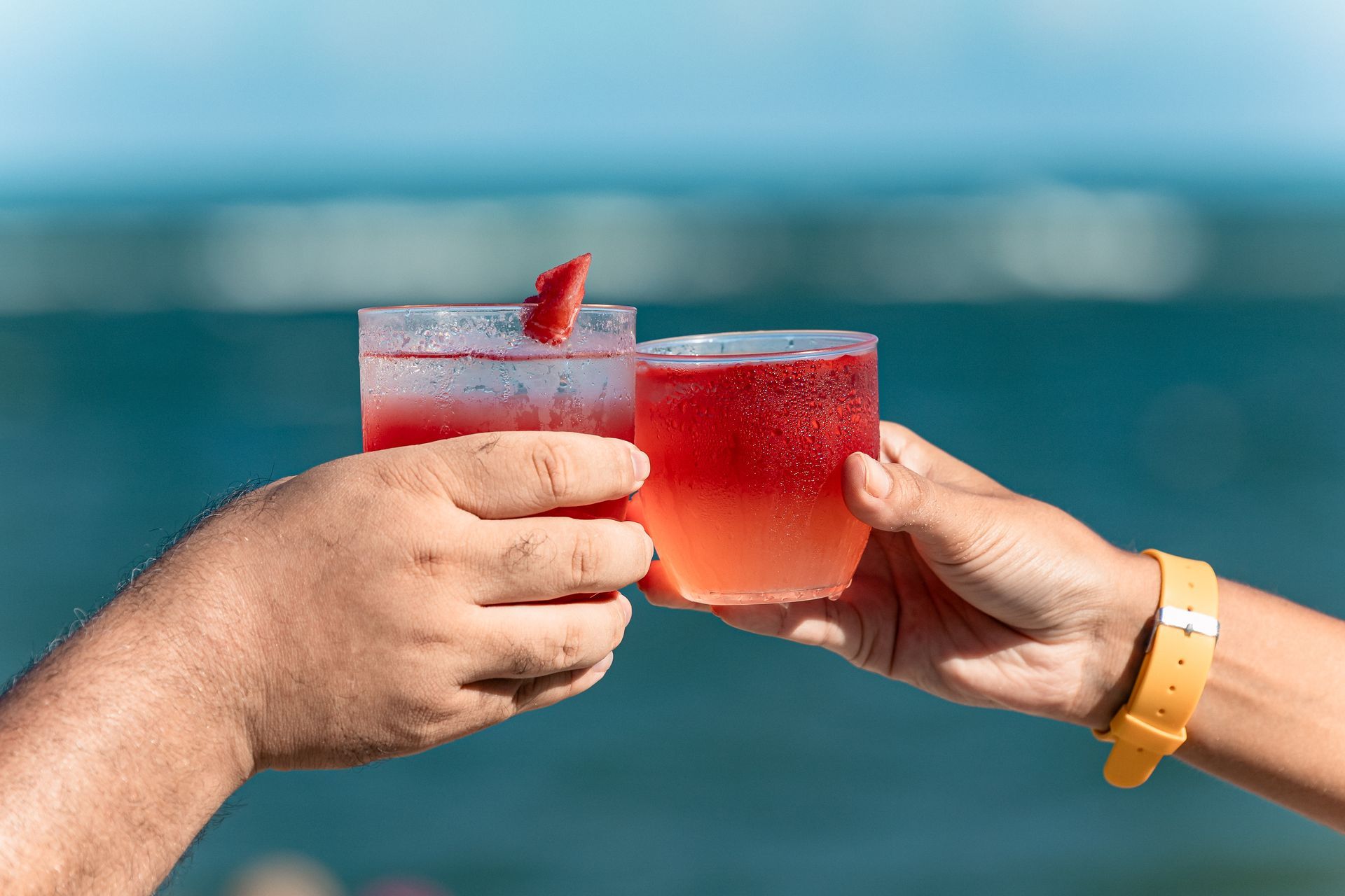 Duas mãos brindando dois drinks vermelhos de frente para o mar da Praia do Francês