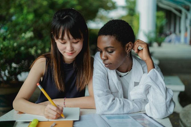 two young adults women sitting next to each other looking down at a piece of paper. one young adult is smiling. 