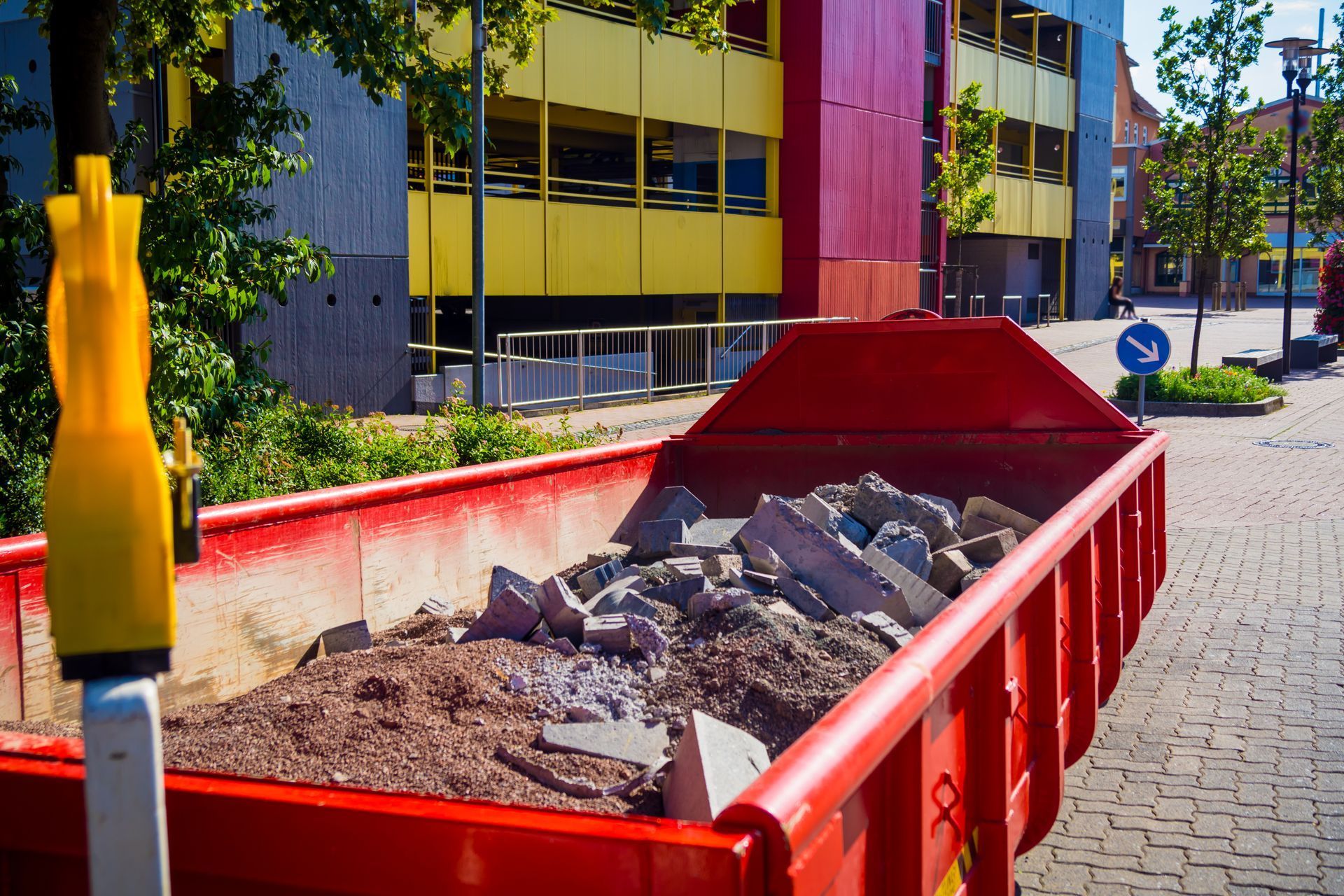 Red dumpster filled with rubble on a paved street. Yellow and red building in the background.