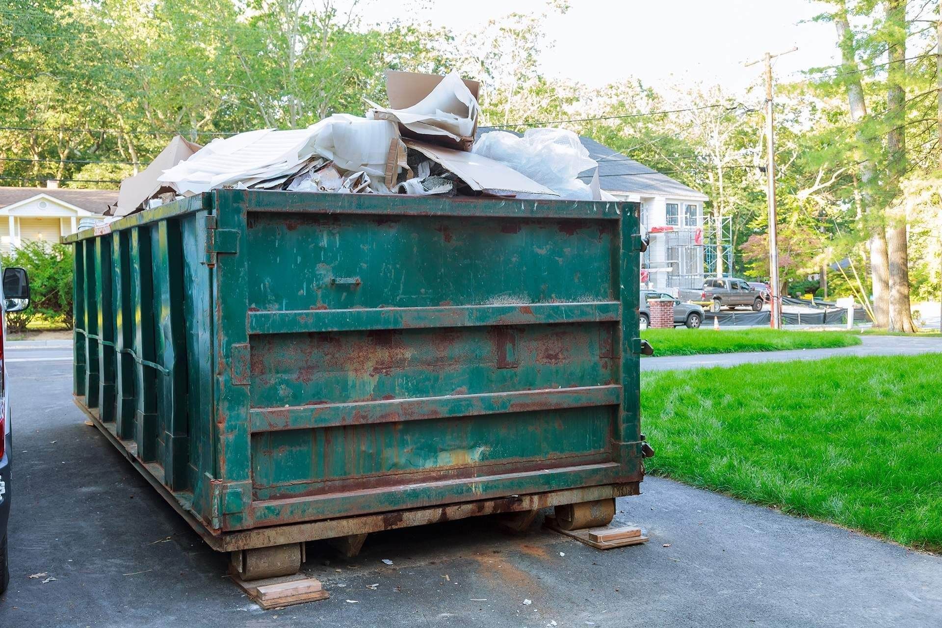Green dumpster overflowing with debris on a driveway, near a residential street.