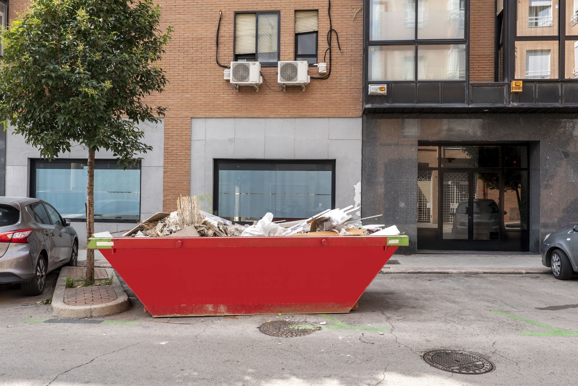 Red dumpster overflowing with debris on a city street, in front of a building.