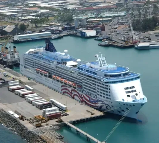 Cruise ship with red, white, and blue design docked in a harbor. Buildings and cranes are in the background.