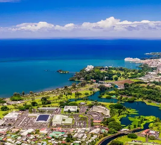 Aerial view of a coastal city with blue ocean, green park, and white buildings under a blue sky.