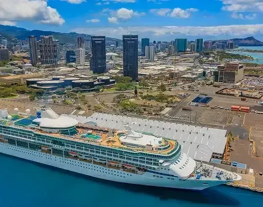 Cruise ship docked at a port in Honolulu, with city buildings in the background and blue water.