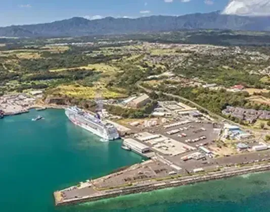 Cruise ship docked in harbor; surrounding buildings and lush green mountains in background.