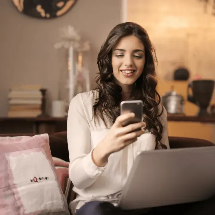 Woman smiling while using a phone and laptop on a couch in a living room.