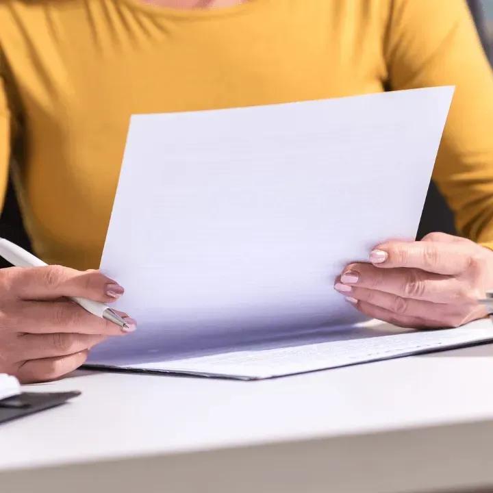 Person in yellow shirt holding a pen and looking at a blank white paper at a desk.