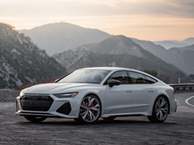 White Audi RS7 parked on a road overlooking mountains at sunset.