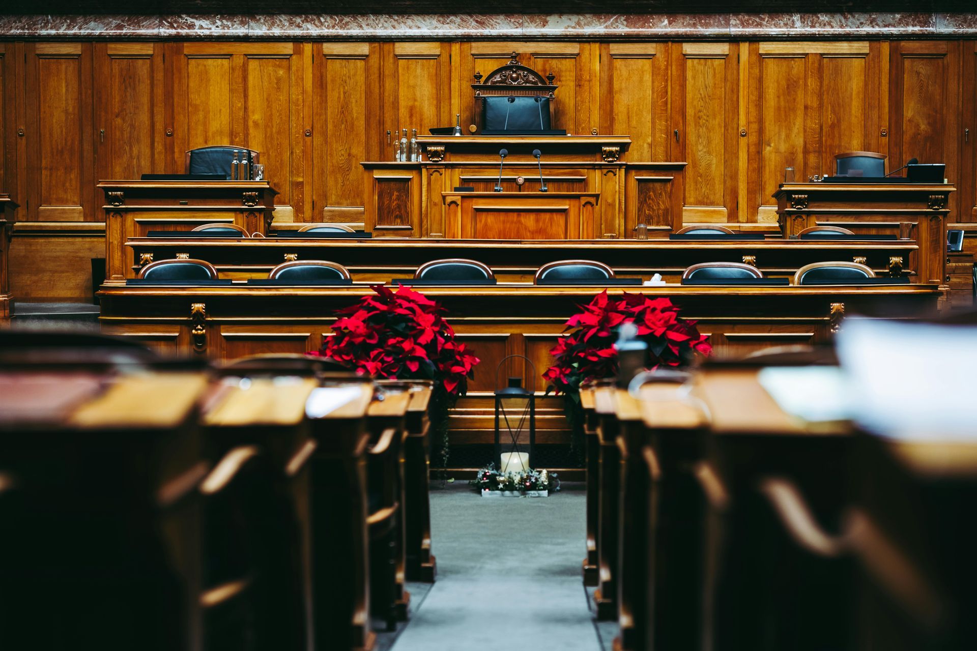 A wood-paneled courtroom with rows of benches, a judge's desk, and red poinsettias.