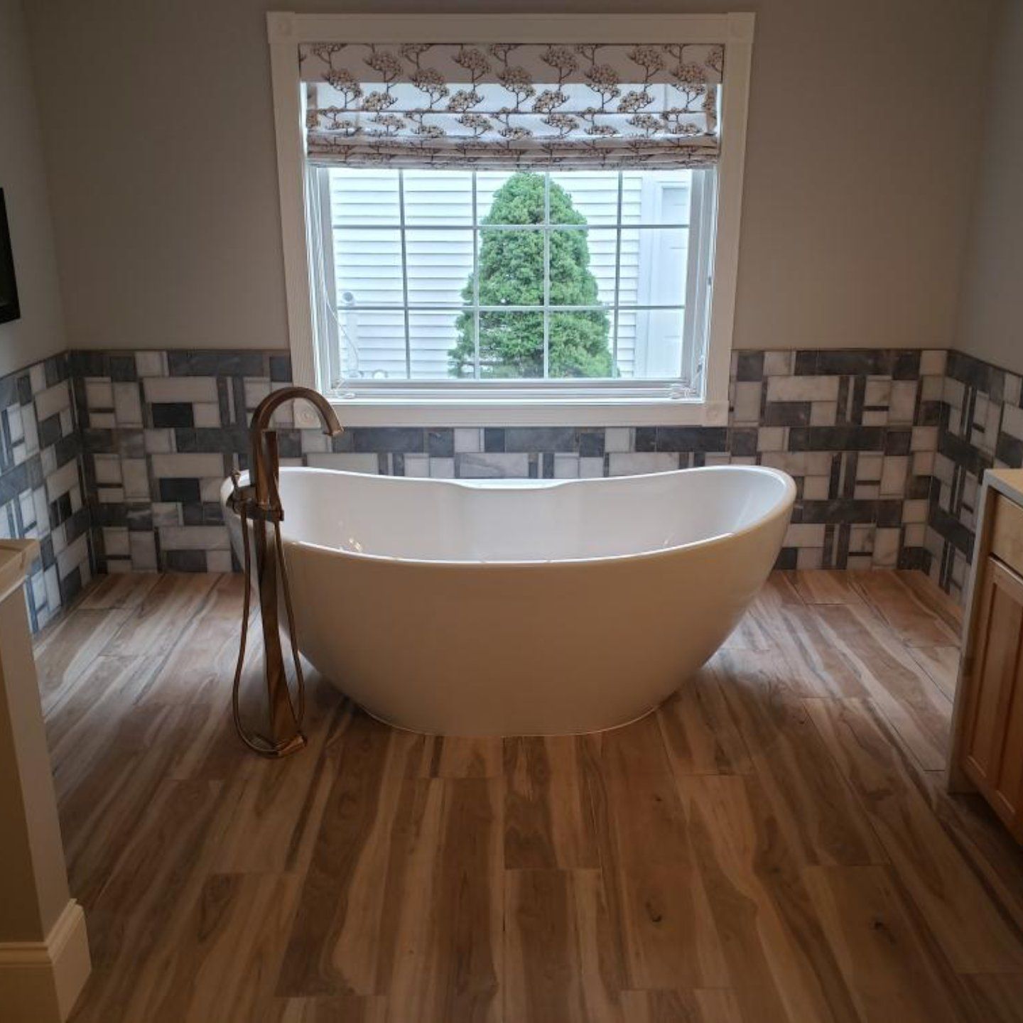 Bathroom with a white oval bathtub, wood-look floor, and patterned wall tile. A window offers a view of green foliage.