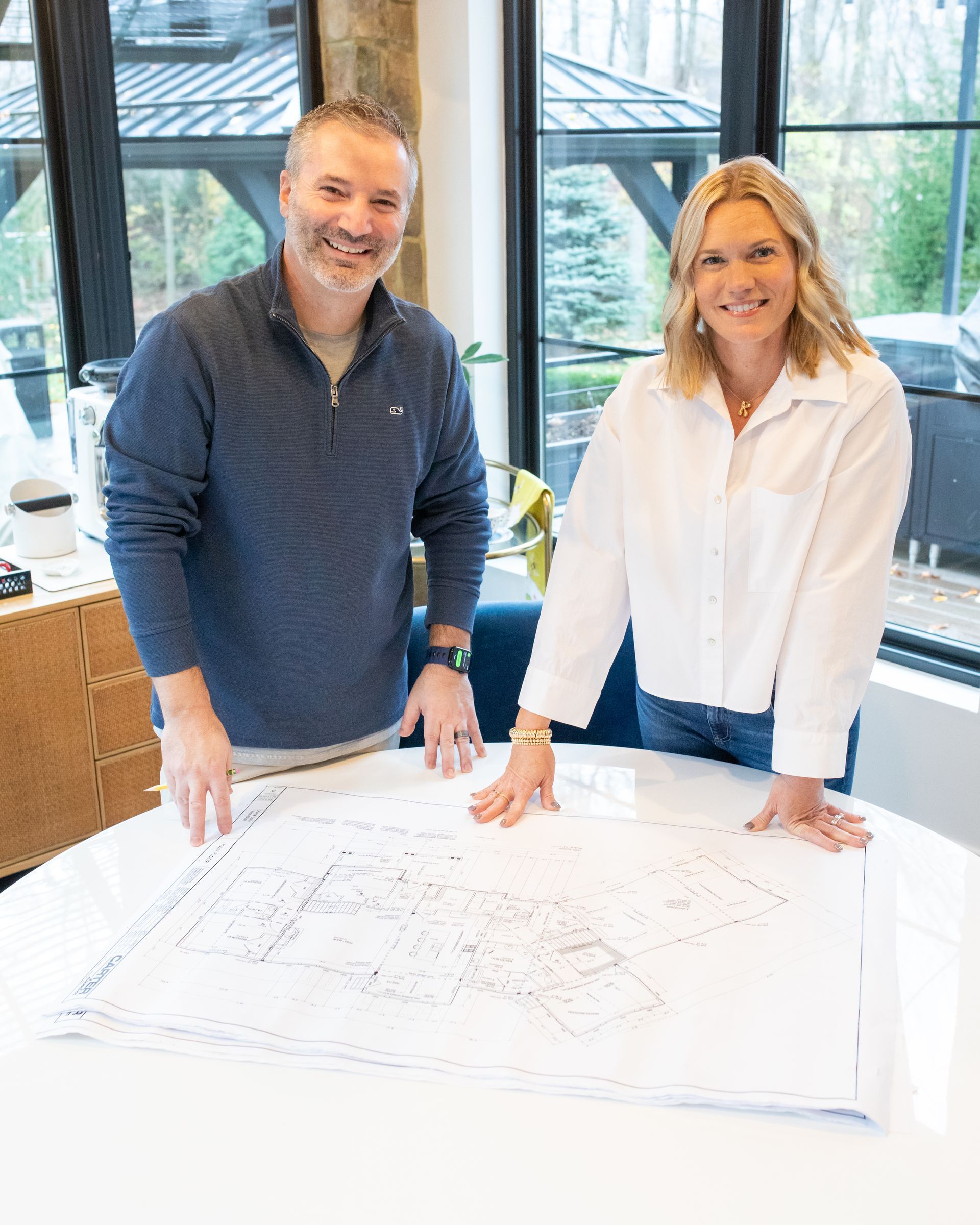 Two people looking at blueprints on a table. Man in blue shirt, woman in white shirt. Bright, modern setting.