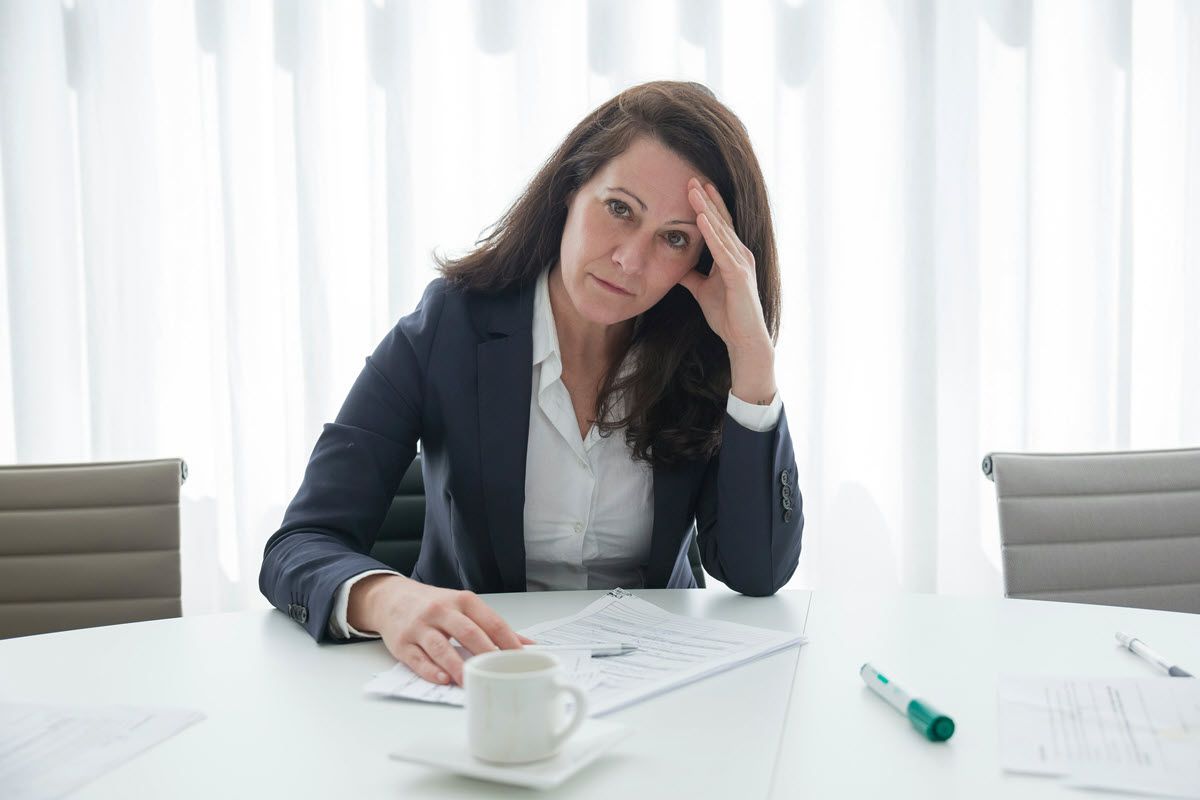 Woman in a blazer at a table, looking stressed with hand on head, papers and coffee mug present.