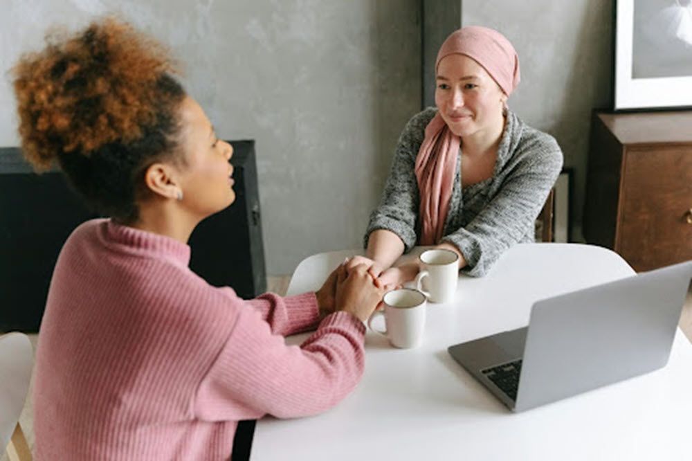 Two women seated at a table, holding hands and smiling. One wears a pink headscarf. Laptop and mugs present.