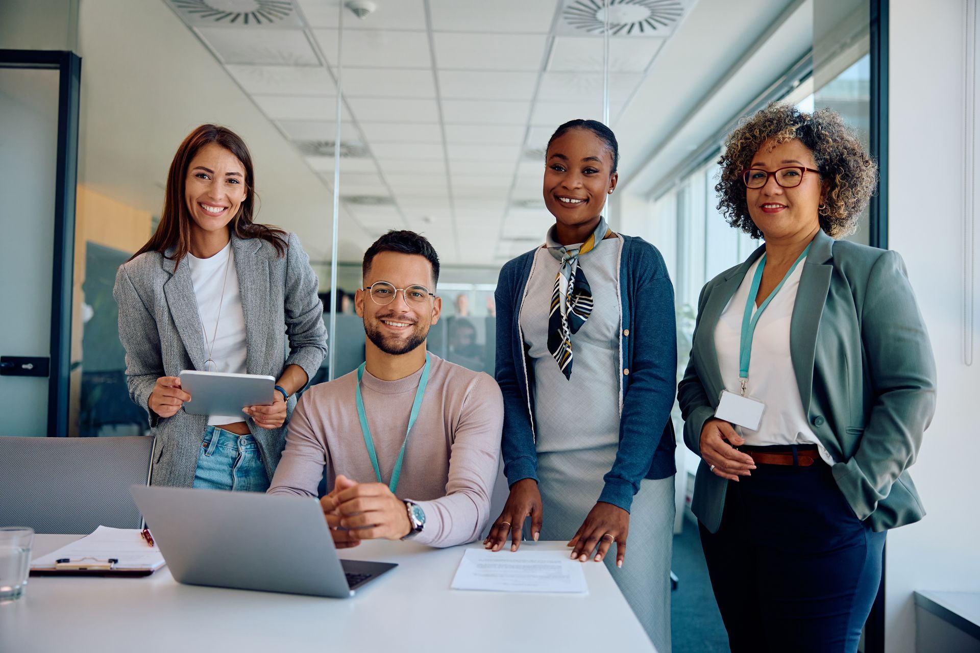 Four coworkers smiling at the camera in an office setting. They are wearing business attire, and one is using a laptop.