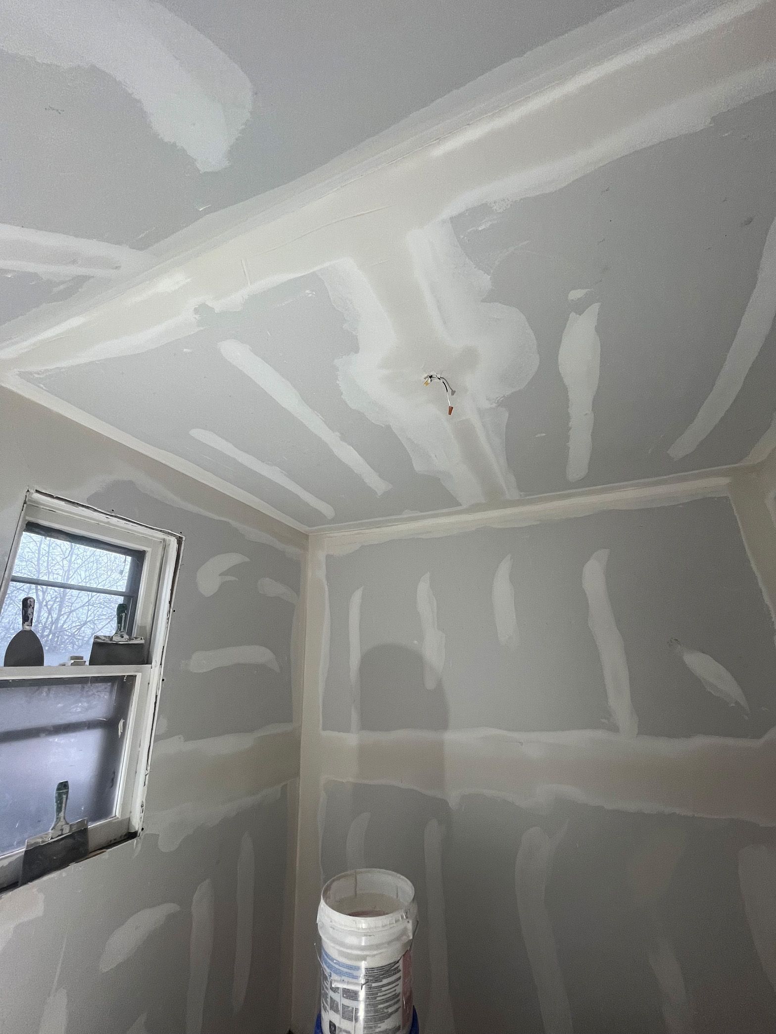 A room with a window and a bucket of paint on the floor.