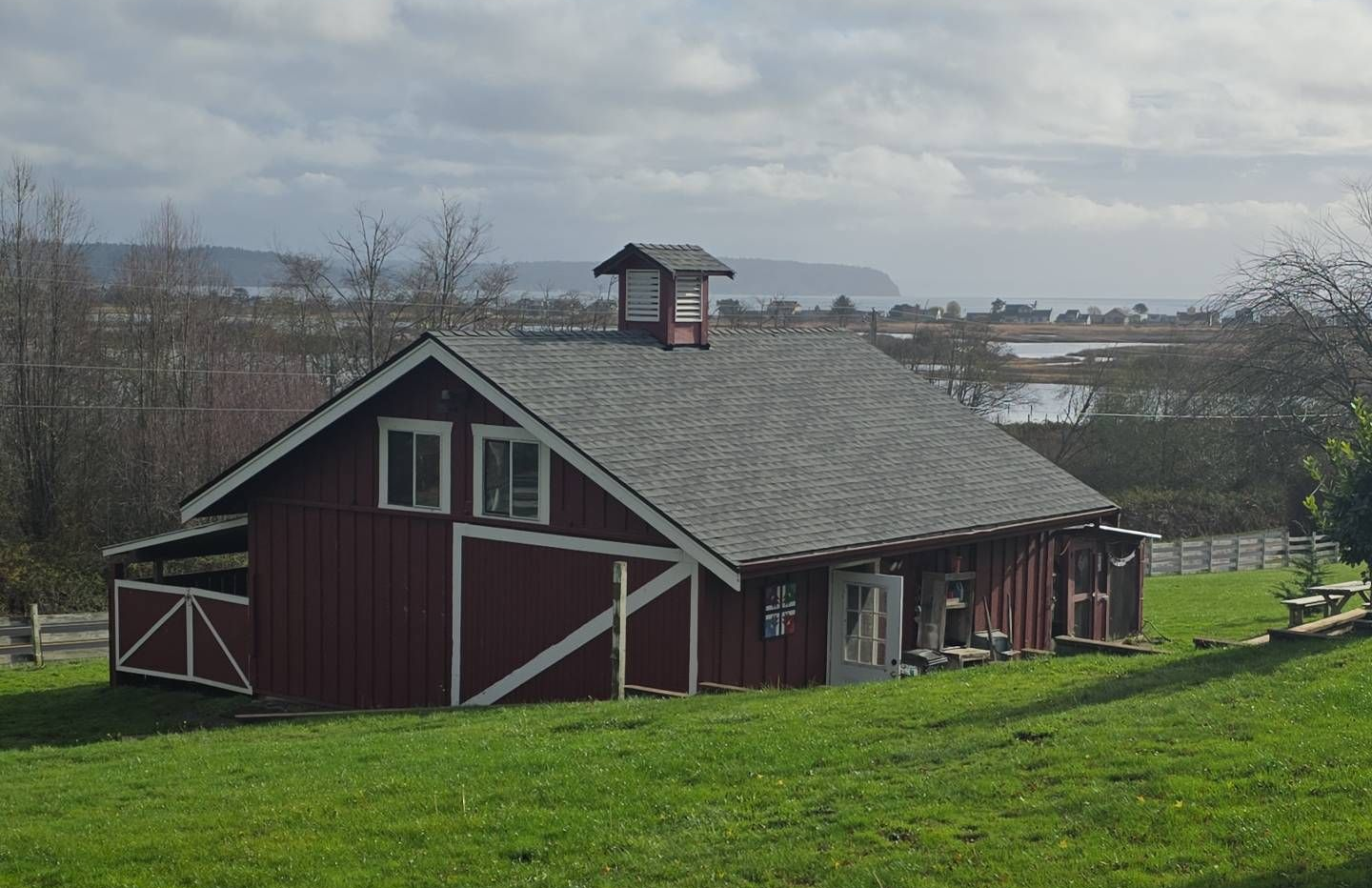Red barn with white trim, set on green grass, overlooking a body of water under a cloudy sky.