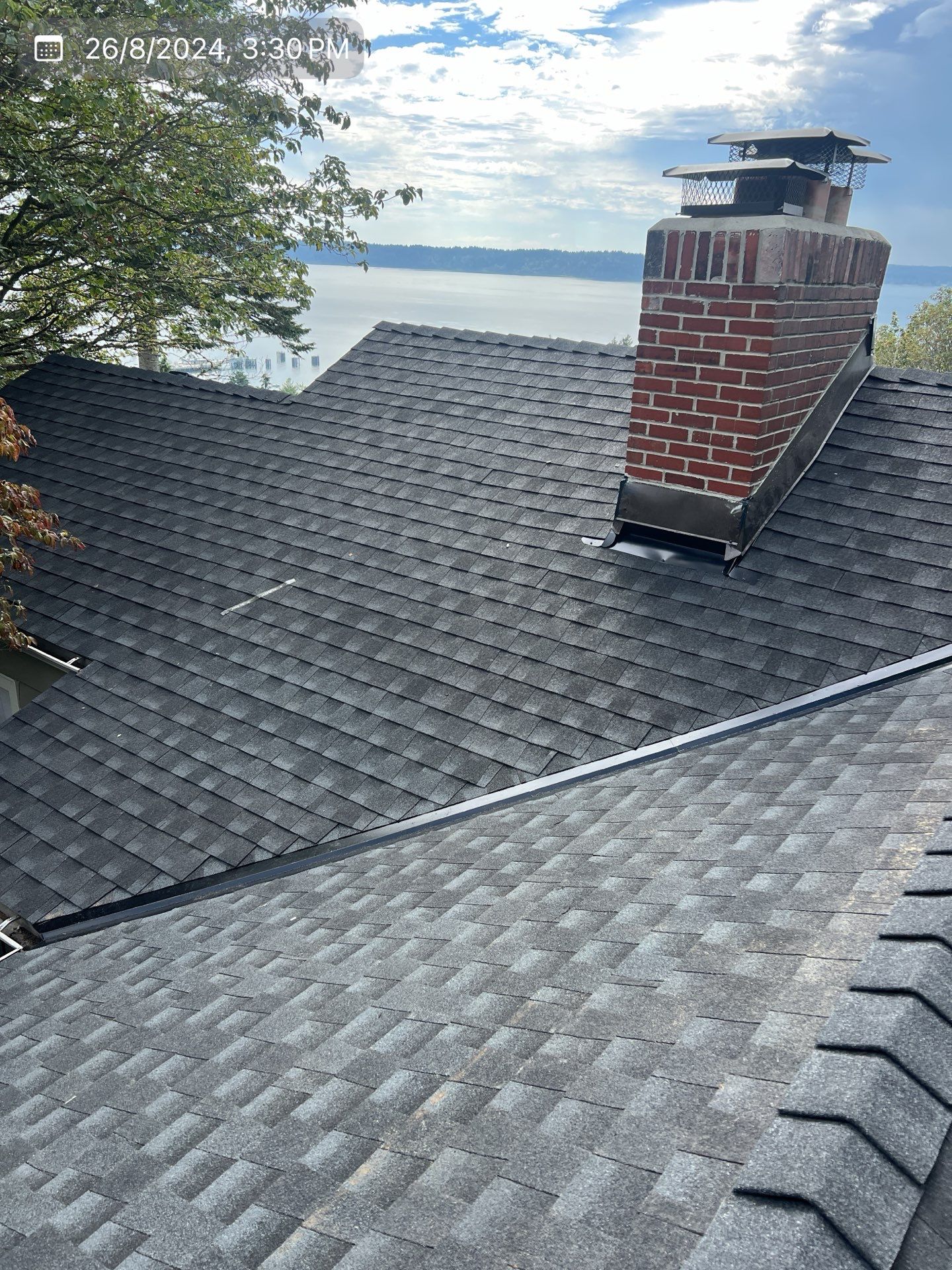 Dark-shingled roof with a brick chimney, overlooking a body of water and trees under a cloudy sky.