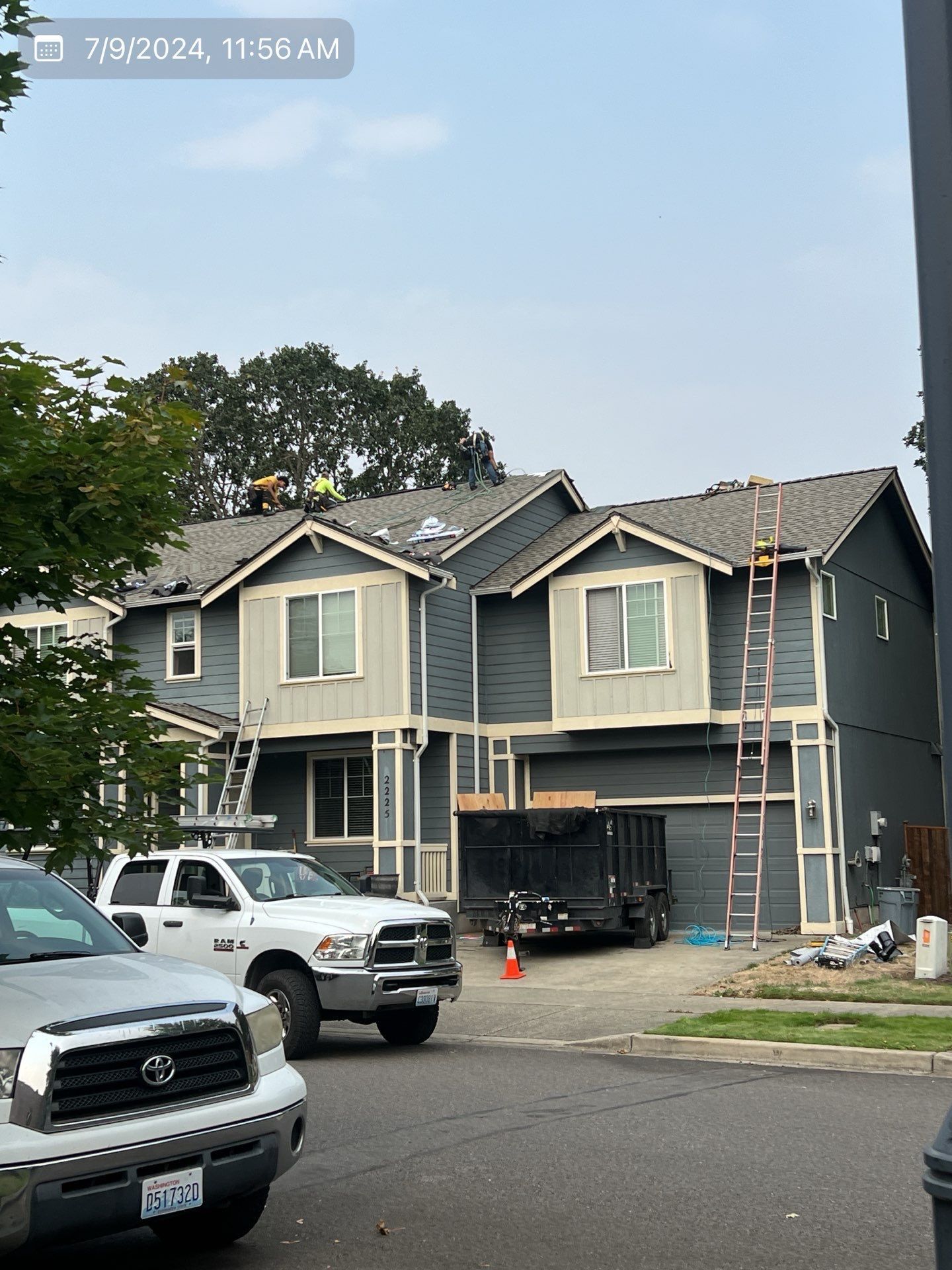 Construction workers on a two-story blue house roof, with a white truck, ladder, and trees.