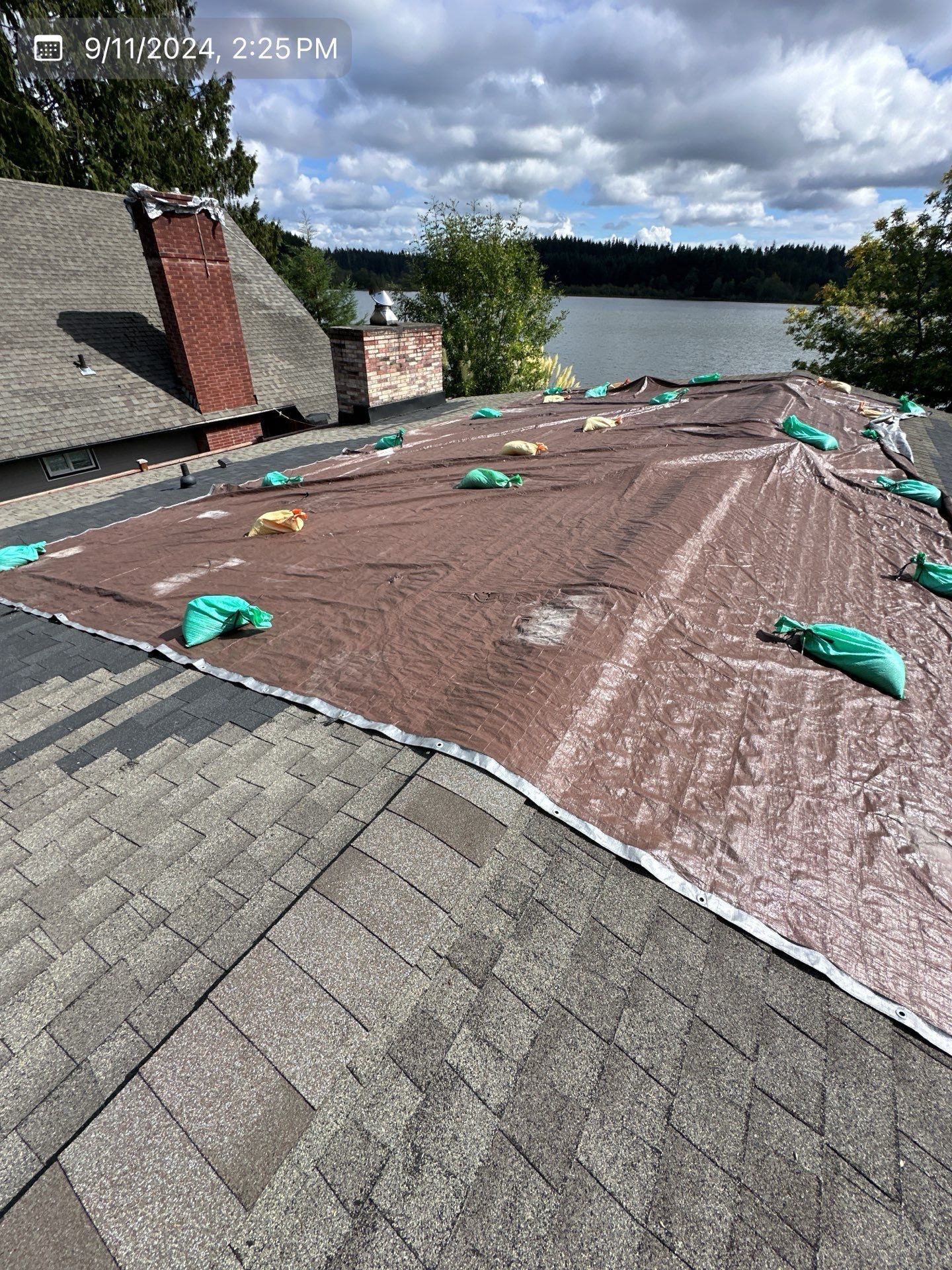 Brown tarp covers a damaged roof, secured with sandbags. Lake and sky in background.