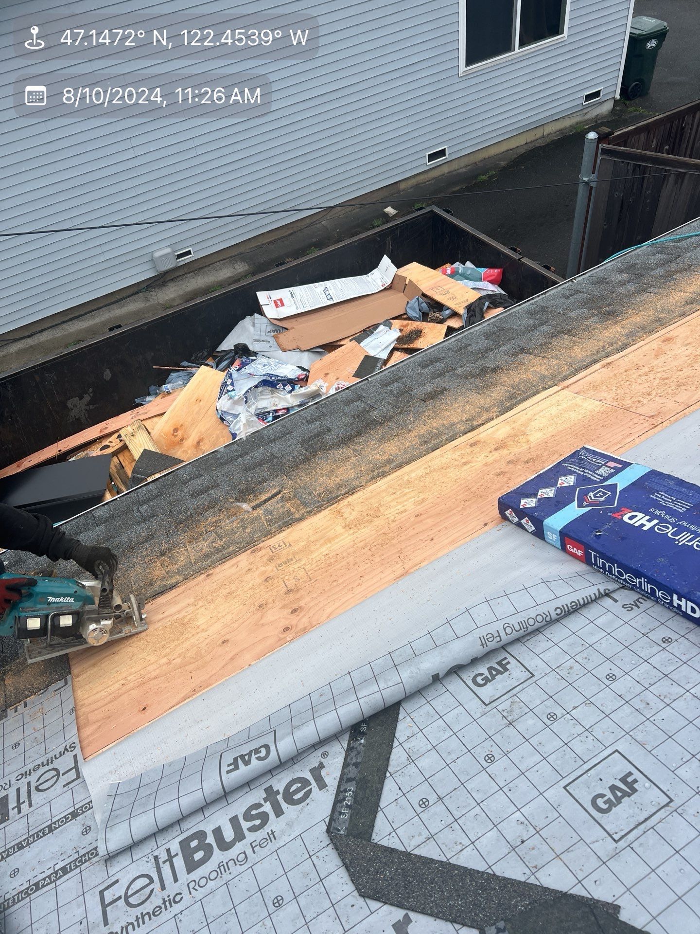 Person using a saw on a rooftop with a dumpster full of debris nearby. FeltBuster roofing materials are in view.
