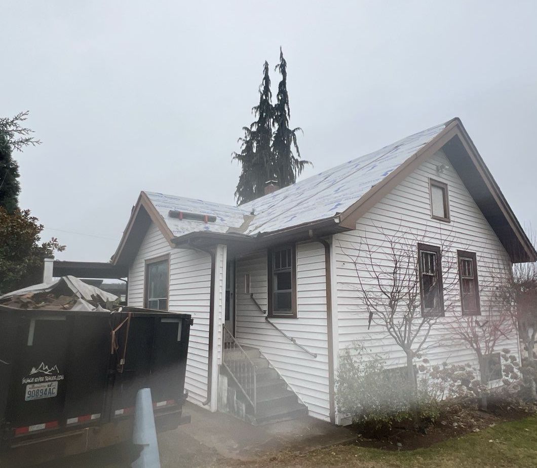 White house with weathered siding; snow on roof, dark dumpster in foreground. Overcast day.