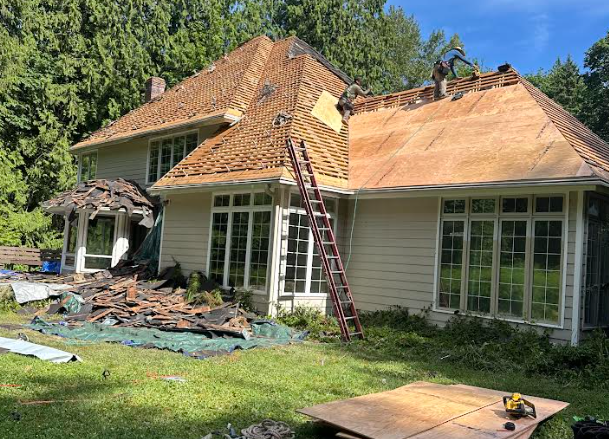 House roof under construction; workers on roof removing shingles, ladder propped, debris on ground.