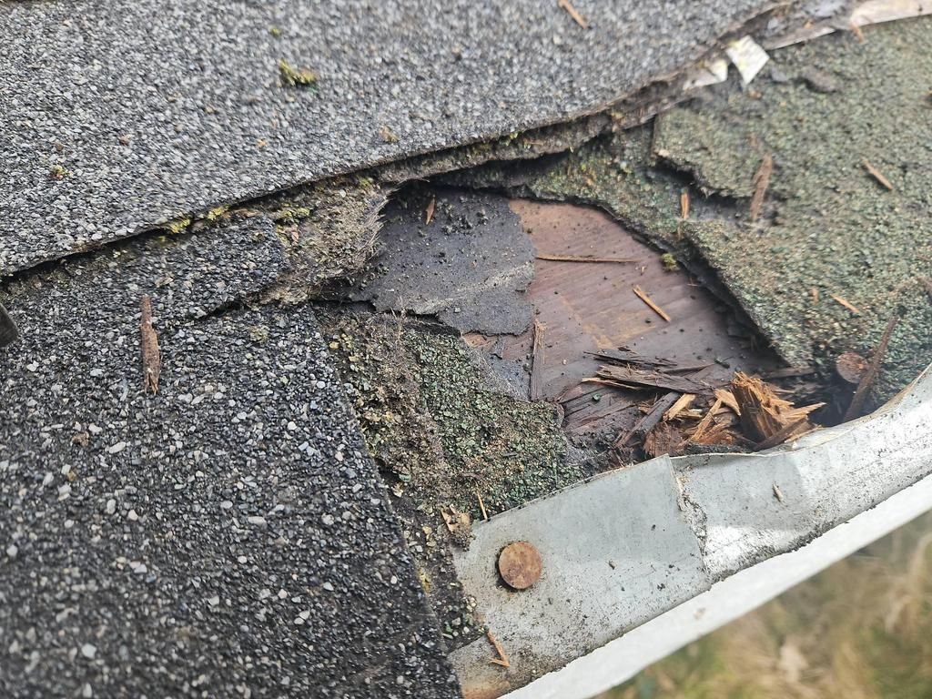 Damaged roof corner with missing shingles and exposed wood, debris in gutter.