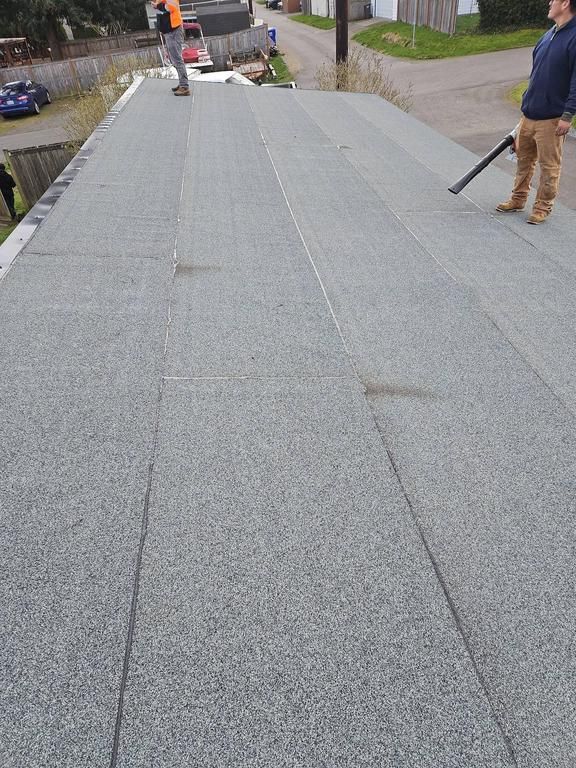 Two people working on a flat, gray, gravel-covered roof; one uses a blower.