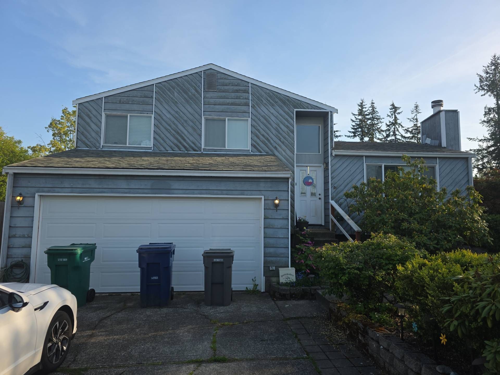 Blue two-story house with a white garage door, trash cans, and a car in the driveway.