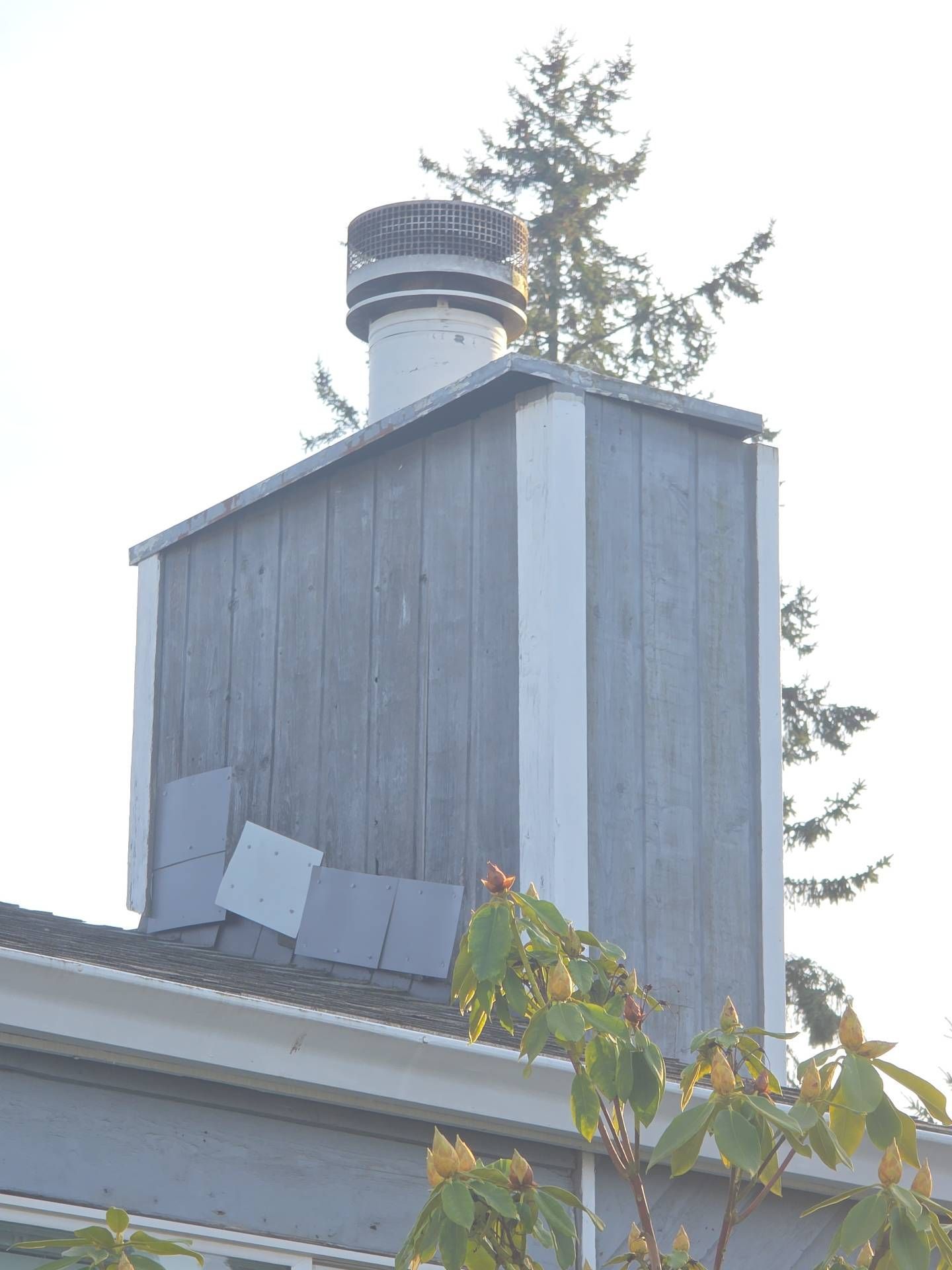 Chimney with weathered gray siding, white trim, and a roof with missing tiles; greenery in foreground.