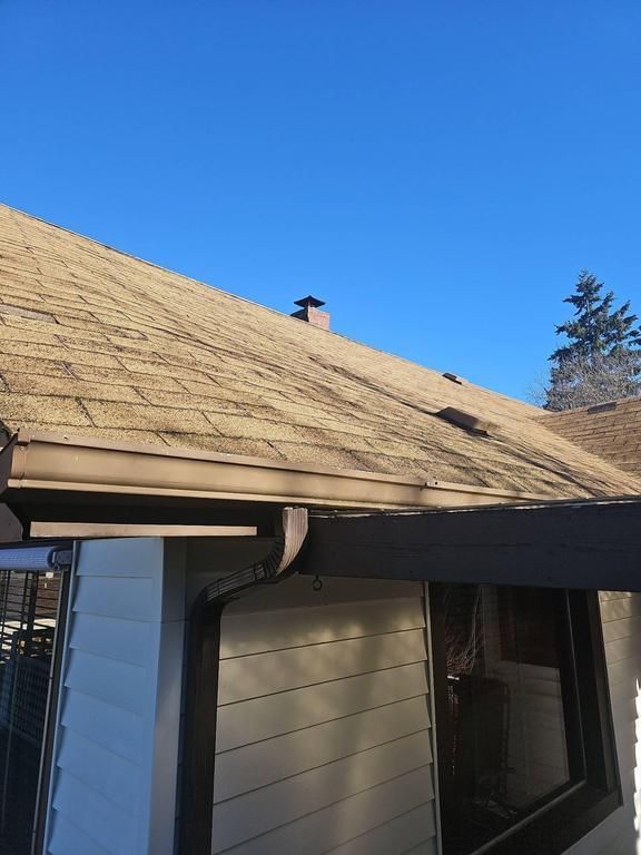Brown shingled roof with a chimney and gutter against a clear blue sky.