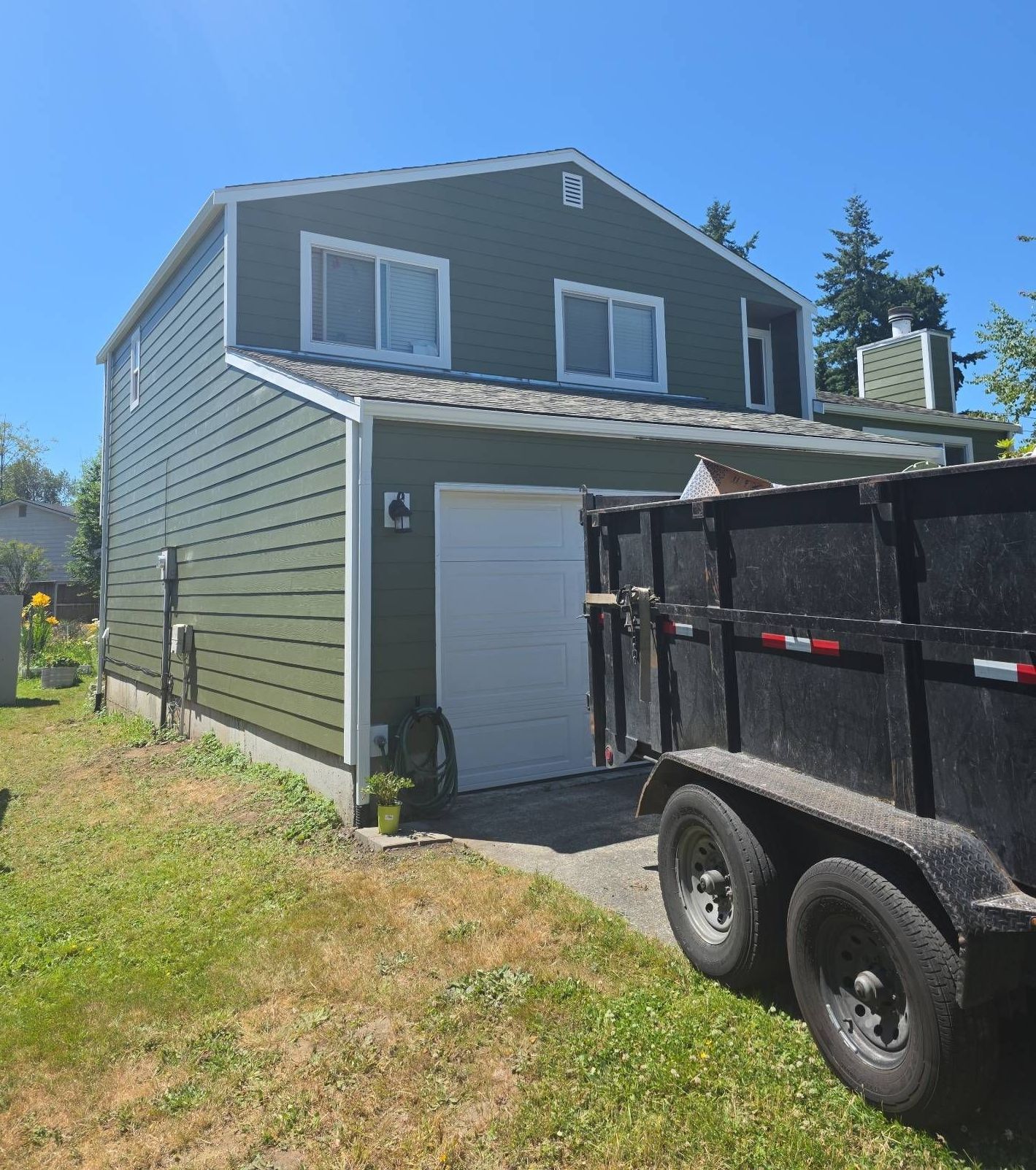 Two-story house with green siding, white trim, and a closed garage door; a trailer is parked nearby.
